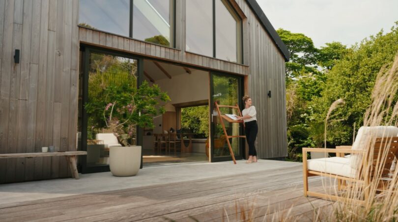 A woman stands at an easel painting on the patio of a modern wooden house with large glass doors, surrounded by greenery and outdoor furniture