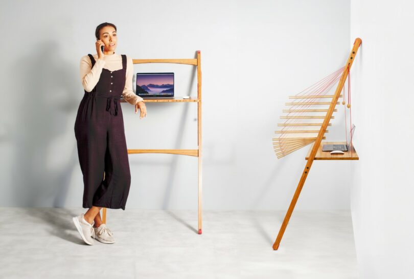A woman stands and talks on the phone next to a wooden standing desk with a laptop, while another wooden structure with shelves is attached to the wall nearby