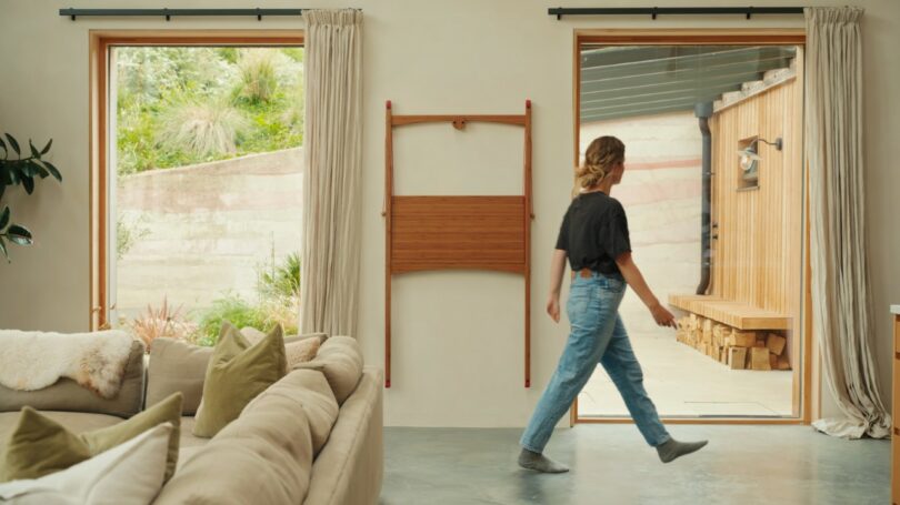 A person in jeans and a black shirt walks through a modern living room with large windows, beige walls, and a beige sofa