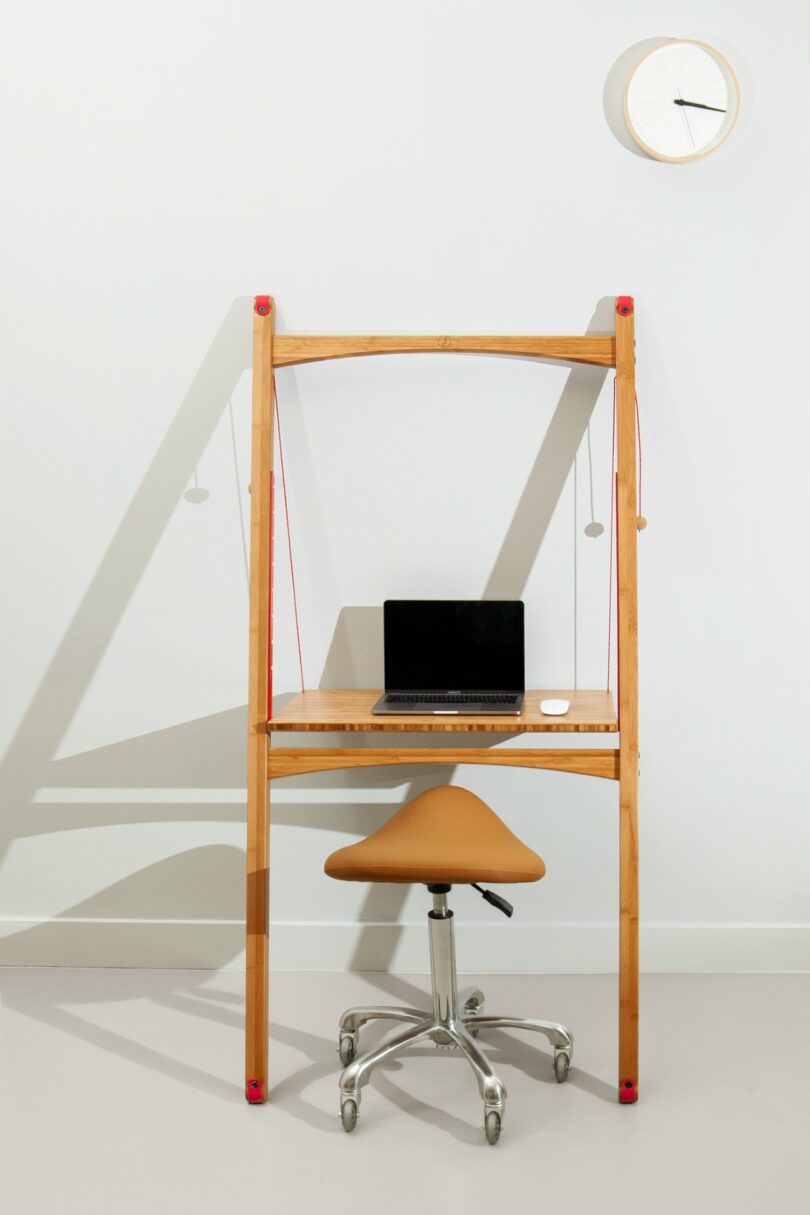 A minimalist wooden desk setup with a closed laptop, wireless mouse, and tan office chair; a wall clock hangs above against a plain white wall