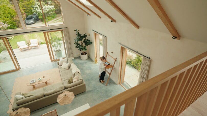A person stands at a wooden desk working on a laptop in a bright, modern living room with large windows overlooking a garden