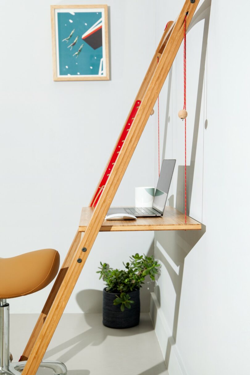 A minimalist workspace with a wooden ladder-style desk, laptop, notebook, tan chair, potted plant, and framed picture on a white wall