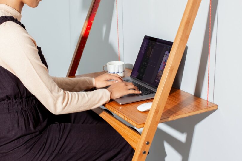 Person sitting at a wooden desk, typing on a laptop with a cup and computer mouse beside them