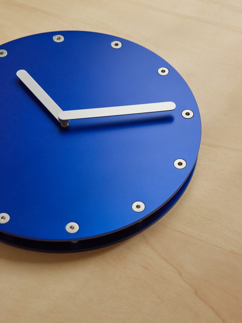A blue wall clock with silver hour and minute hands is mounted on a light wood surface, showing the time 