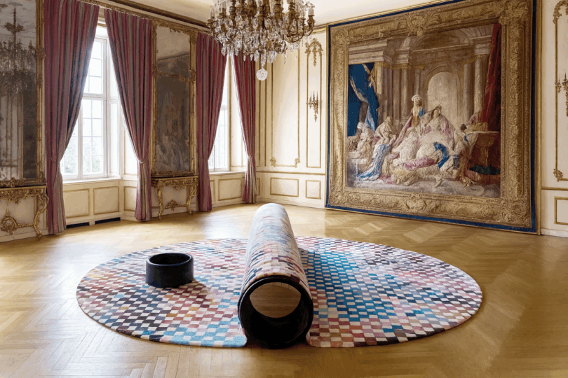 bench-carpet hybrid on a parquet floor in an ornate room with a chandelier, large tapestry, and tall windows with red curtains.