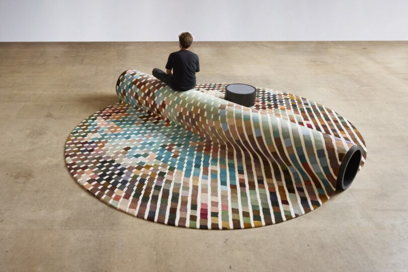 A person sits on a bench-carpet hybrid, with a small round table nearby in a spacious room