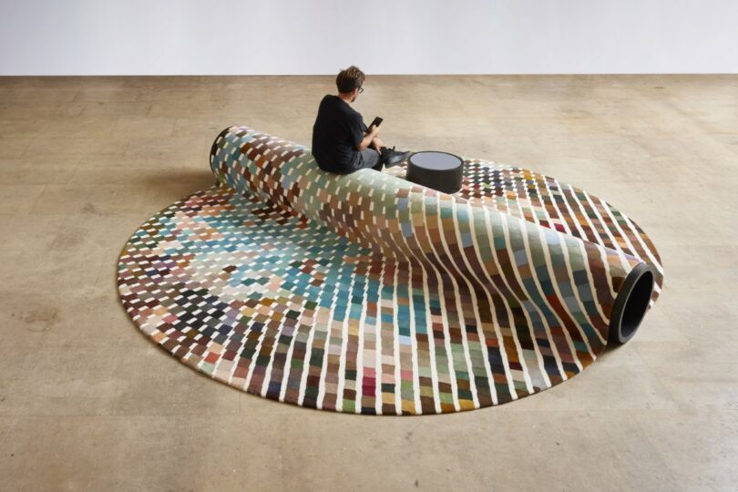 A person sits on a bench-carpet hybrid, with a small round table nearby in a spacious room