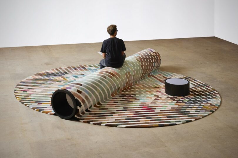 A person sits on a bench-carpet hybrid, with a small round table nearby in a spacious room