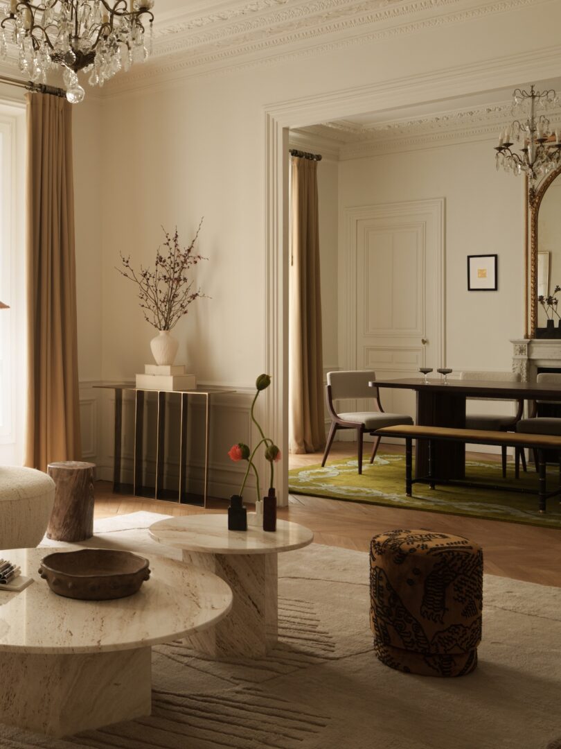 Elegant living and dining room with beige walls, marble tables, modern decor, large windows with brown curtains, and ornate ceiling molding.