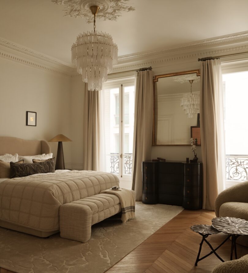 Elegant bedroom with a large upholstered bed, chandelier, ornate ceiling, tall windows with beige curtains, a black dresser, mirror, and light wood floors with a beige rug.