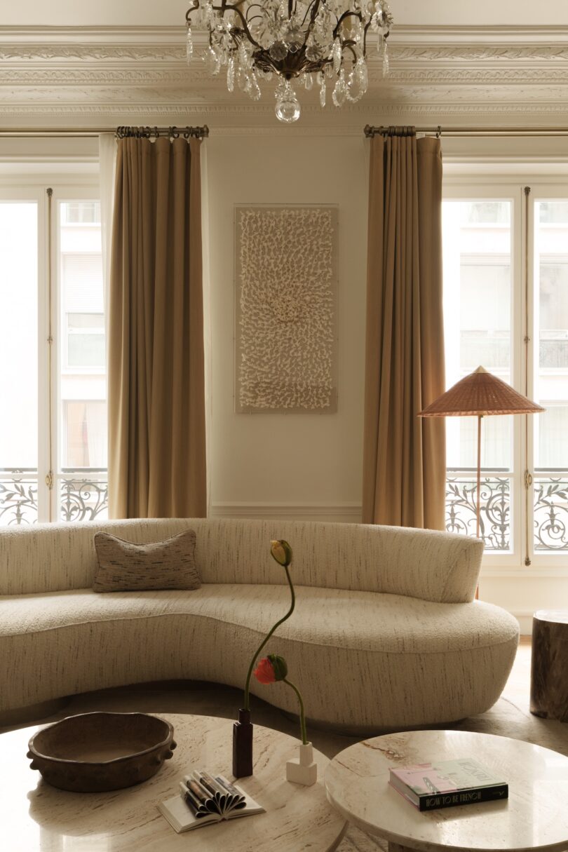 A neutral-toned living room with a curved sofa, round coffee table, decorative bowl, book, single-stem flower, large windows, beige curtains, and a chandelier overhead.