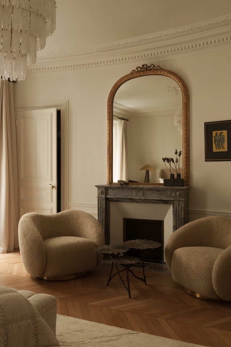 A living room with two beige boucle armchairs, a marble fireplace, a large gold-framed mirror, and a sculptural coffee table on herringbone wood floors.