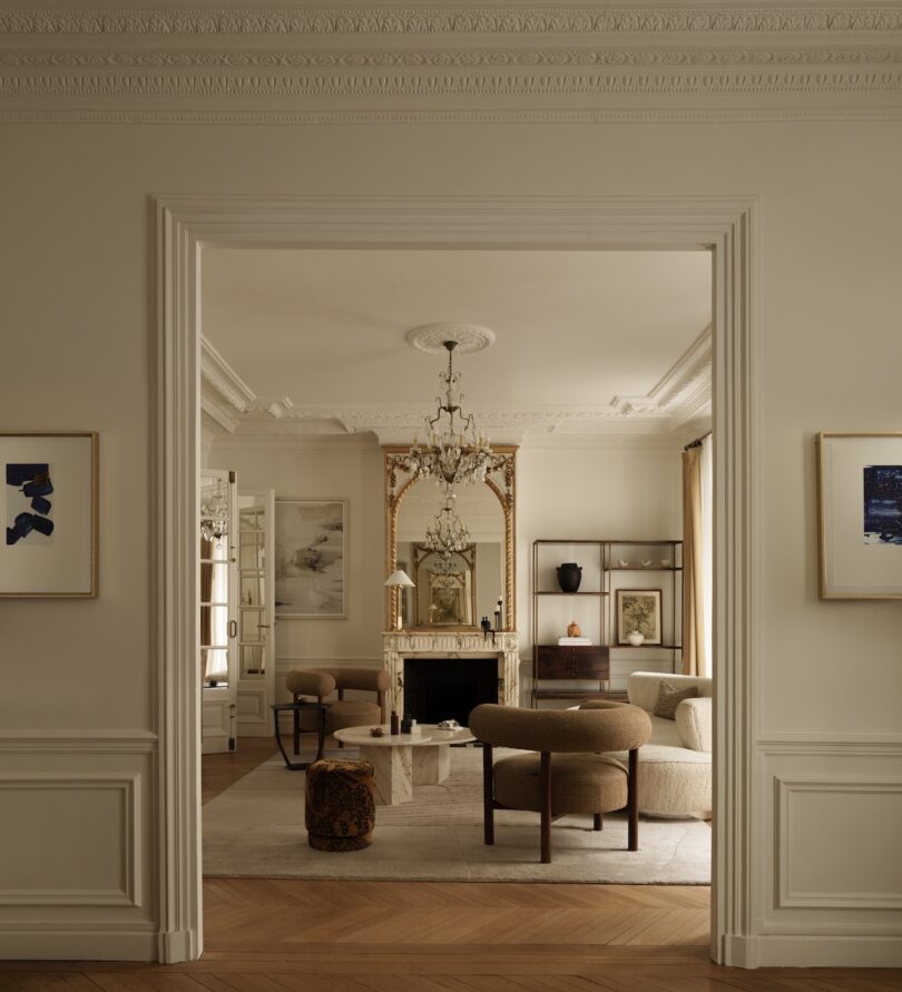 A neutral-toned living room with ornate molding, a chandelier, modern armchairs, a marble fireplace, large mirror, and contemporary artwork on the walls.