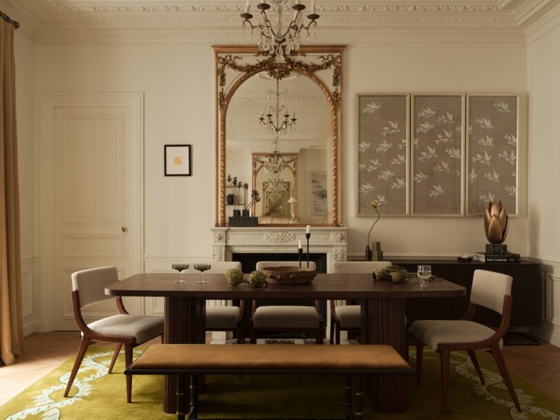 Elegant dining room with a wooden table, chairs, a bench, a large ornate mirror above a fireplace, and wall art with bird motifs. A chandelier hangs from the decorative ceiling.