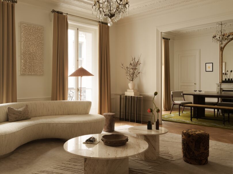 A neutral-toned living room with a curved sofa, marble coffee tables, floor-to-ceiling curtains, a chandelier, and a dining area with a large mirror visible in the background.