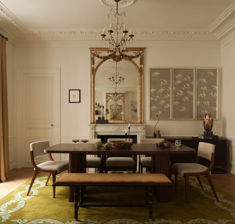 Elegant dining room with a wooden table, chairs, and bench on a green rug. A large ornate mirror and artwork hang above a white fireplace; chandelier overhead.