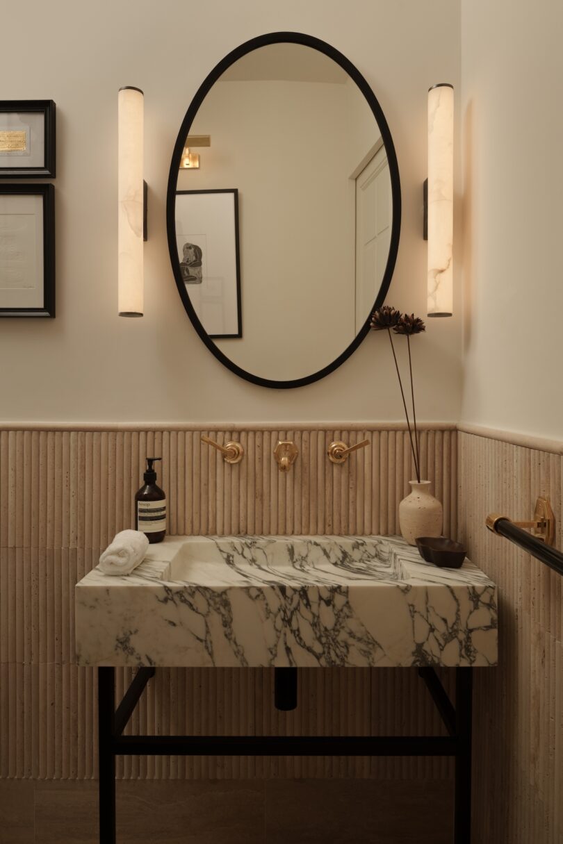 A modern bathroom features a marble sink, oval mirror, wall-mounted faucets, vertical wall lights, decorative vase, and framed art on textured beige walls.