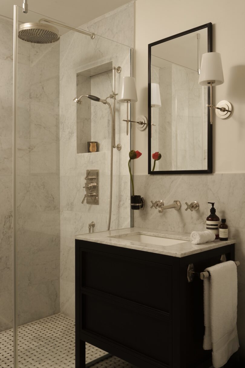Modern bathroom with a glass-enclosed shower, marble walls, black vanity with a marble countertop, wall-mounted faucets, and a large mirror flanked by two wall sconces.