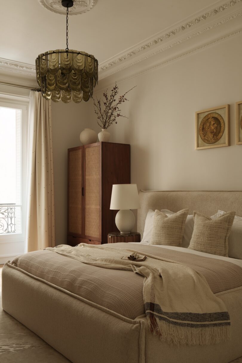 Neutral-toned bedroom with a large bed, wooden wardrobe, bedside table, lamp, wall art, chandelier, and a window with curtains letting in natural light.