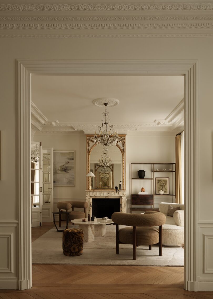 A sophisticated living room with a large mirror above a fireplace, modern beige seating, a marble coffee table, and decorative molding on the walls and ceiling.