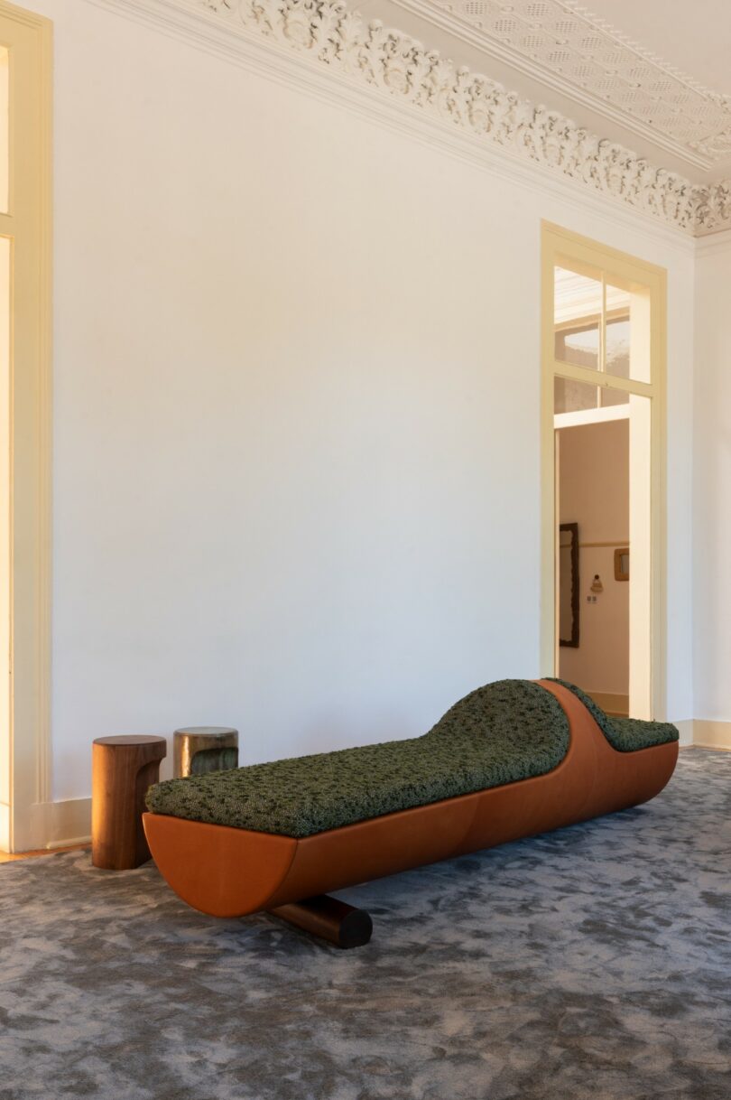 A modern chaise lounge with a green patterned cushion and brown leather sides sits on a gray carpet in a room with ornate ceiling molding and tall doorways