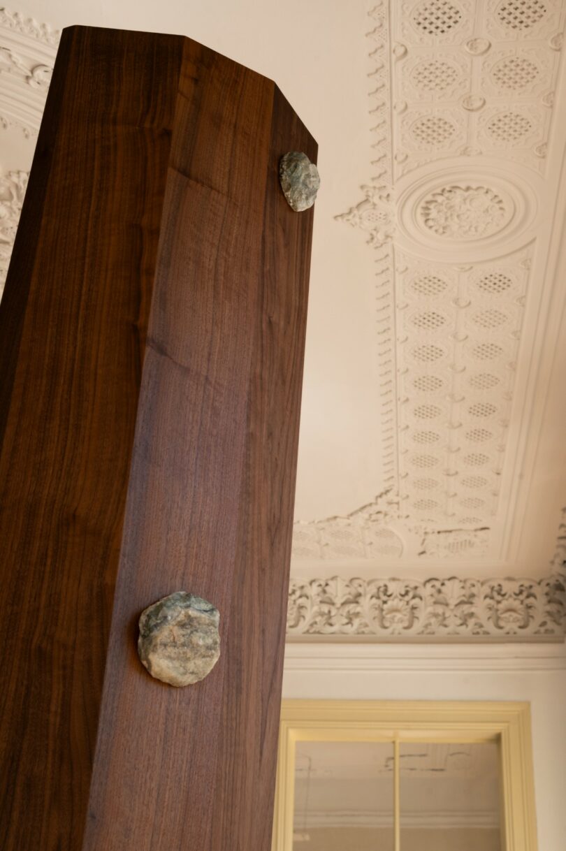 A tall, polished wooden beam with two embedded stones stands in front of an ornate, white, decorative ceiling