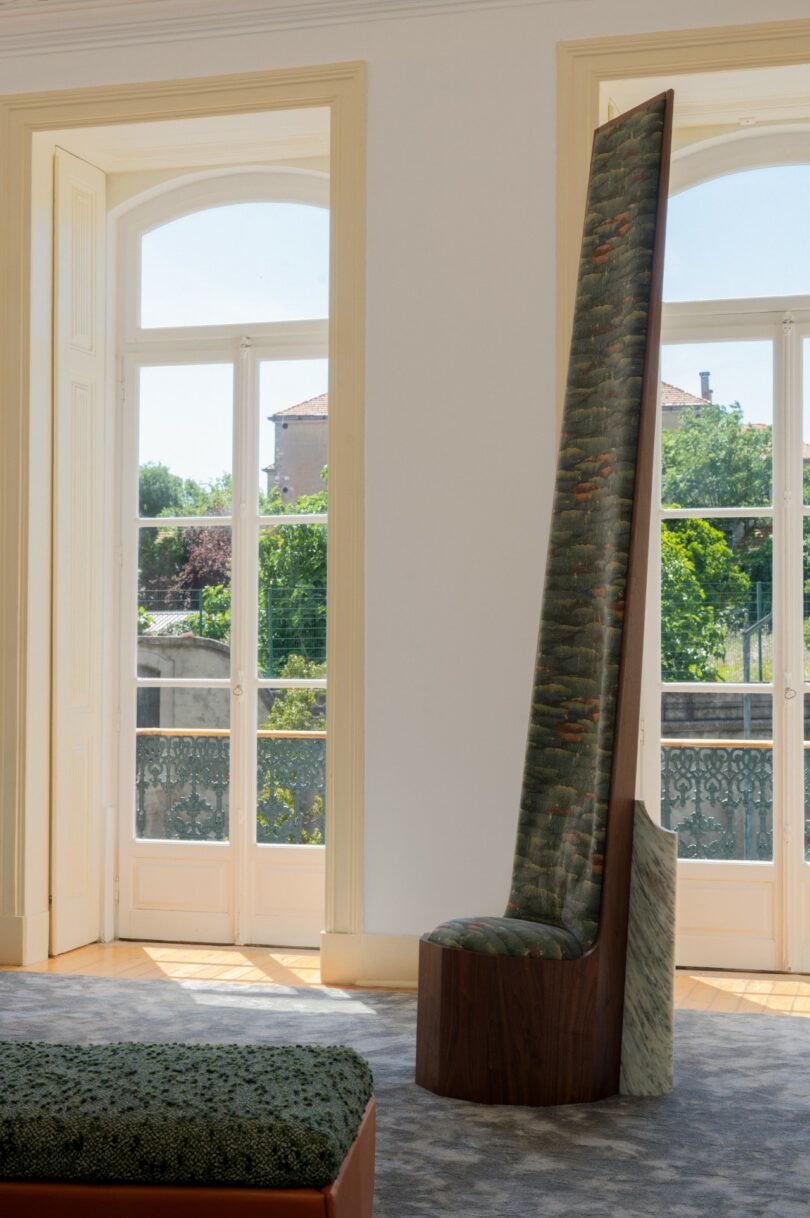A tall-backed chair with green patterned upholstery and a wooden base is positioned in front of large windows in a sunlit room