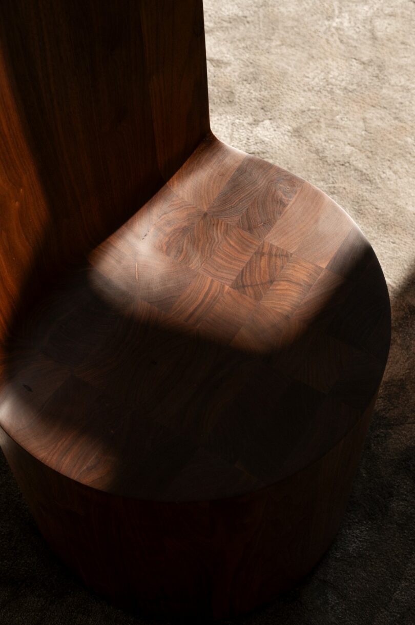 A curved wooden chair seat with a smooth finish is partially illuminated by sunlight, casting a shadow on a textured carpeted floor