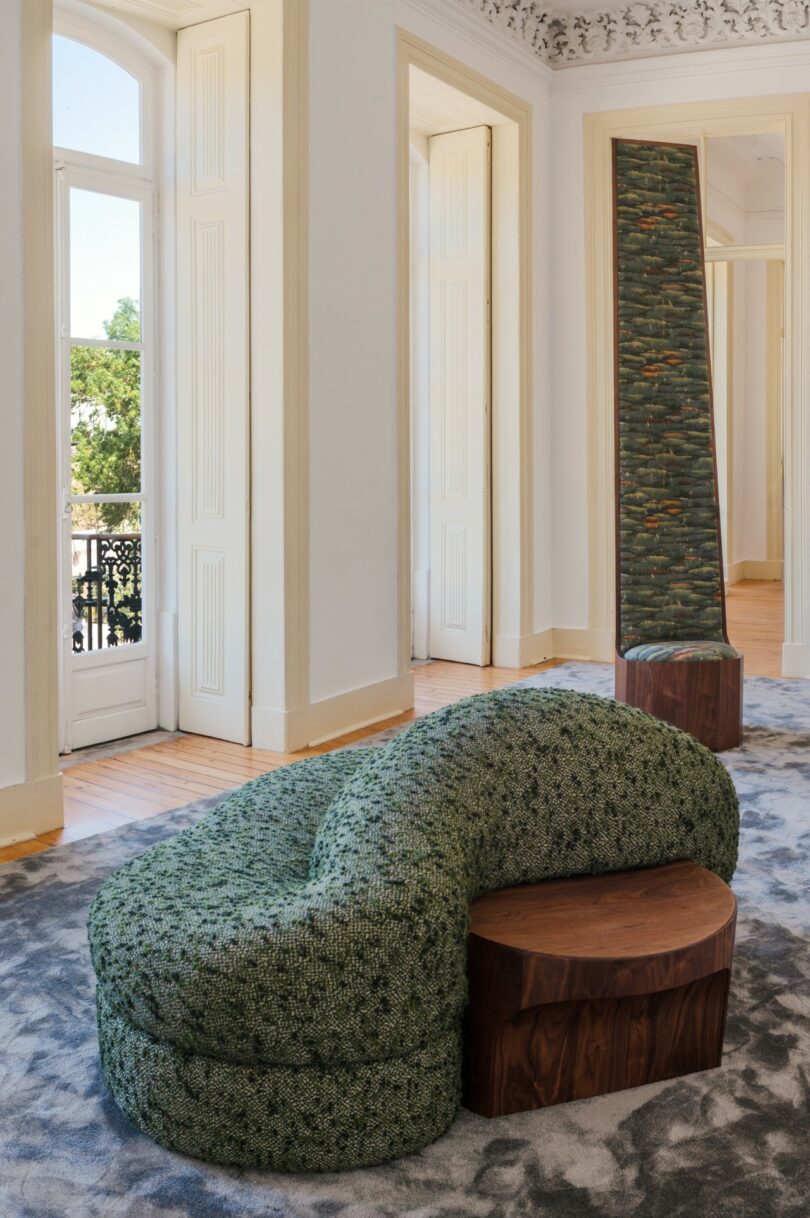A modern room with tall windows features a curved green upholstered sofa with a wooden base and a tall, narrow mirror beside it, both set on a patterned gray carpet