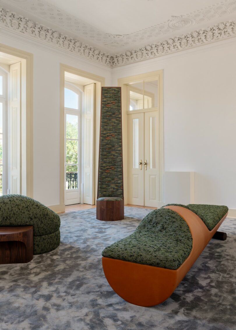 A modern room with ornate ceiling moldings, large windows, and minimalist furniture, including green-cushioned benches and a tall, textured column