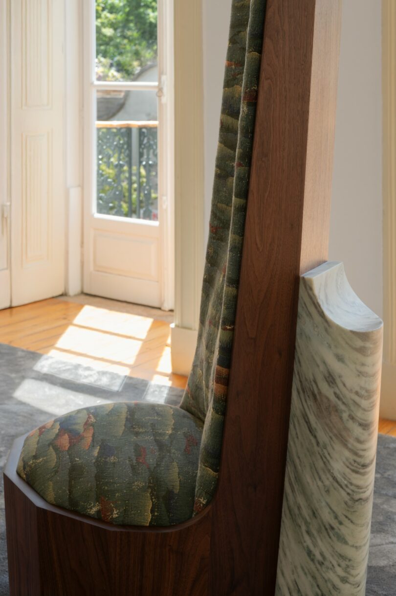 A modern chair with a high wooden backrest, patterned green upholstery, and a marble accent sits in a sunlit room near an open door to a balcony.