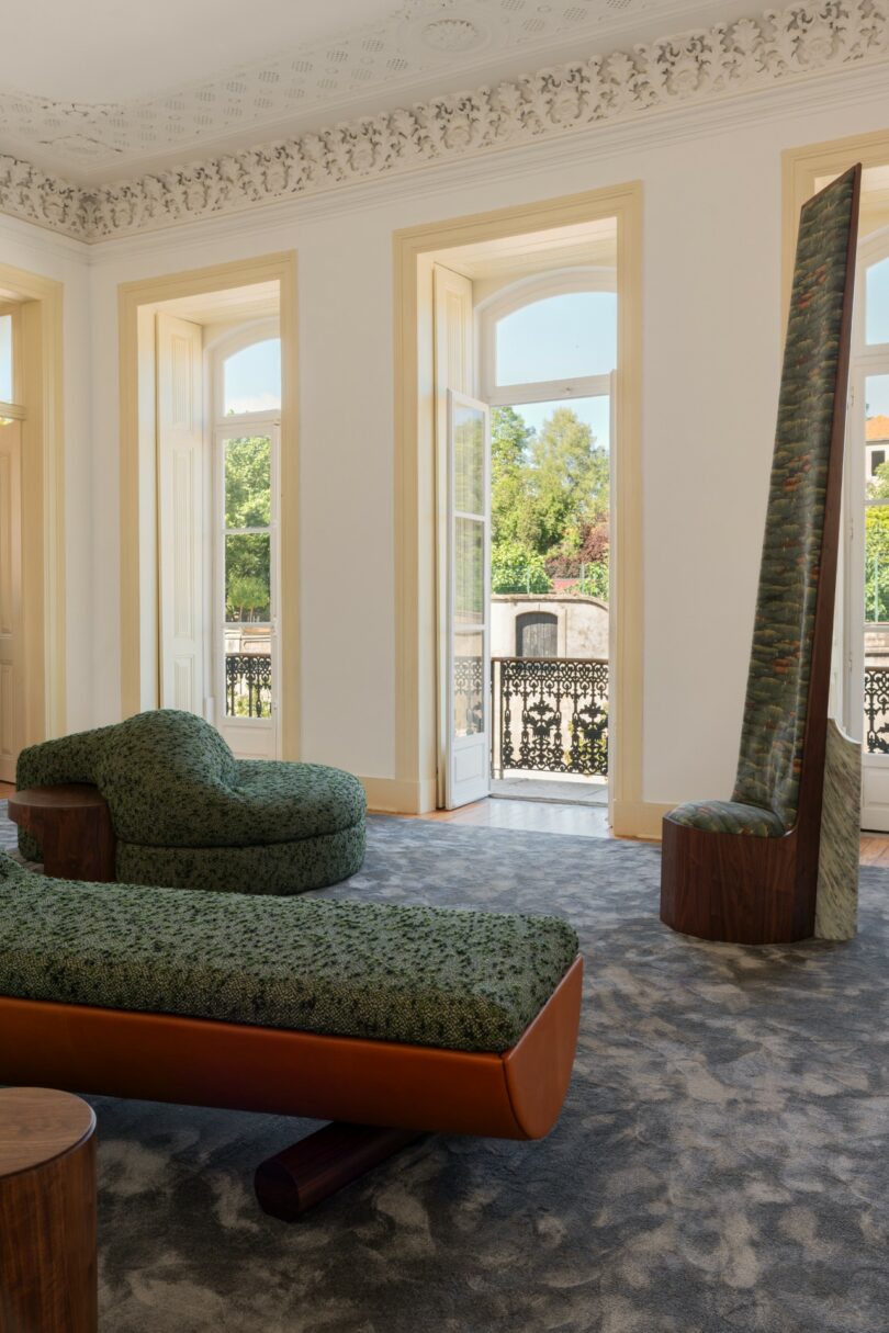 A modern lounge with patterned green furniture, large windows, ornate ceilings, and a balcony overlooking greenery