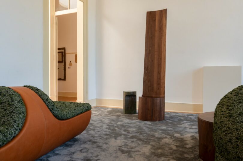 Minimalist room with textured green and brown furniture, a tall wooden sculpture, gray carpet, and white walls with yellow trim