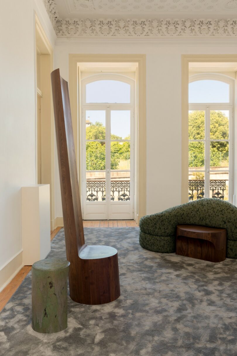 A modern room with tall windows, a high-backed wooden chair, a small green stool, a textured green lounge chair, and a patterned gray carpet