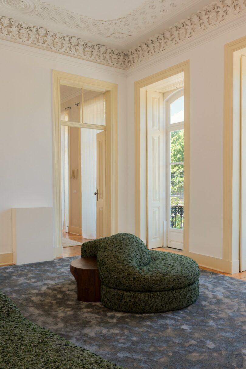 A modern living room with high ceilings, decorative molding, large windows, a green patterned chaise lounge, a wooden side table, and a blue-grey rug