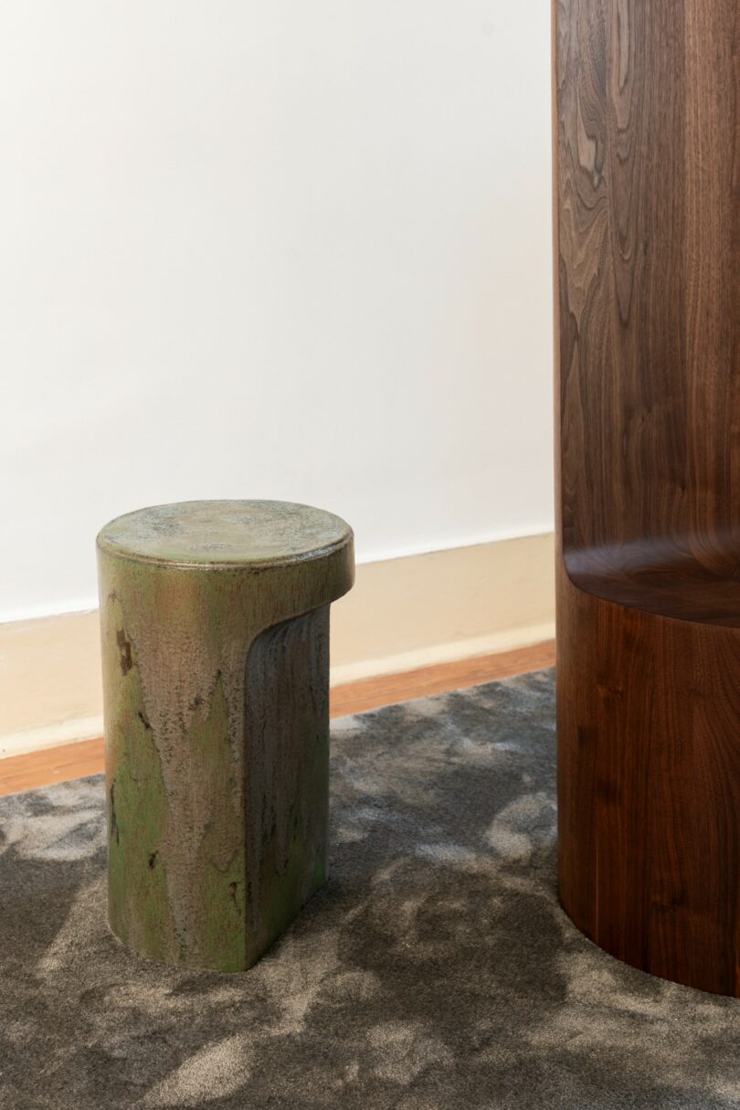 A small, moss-colored stone stool sits on a patterned carpet next to a curved, dark wooden structure against a white wall