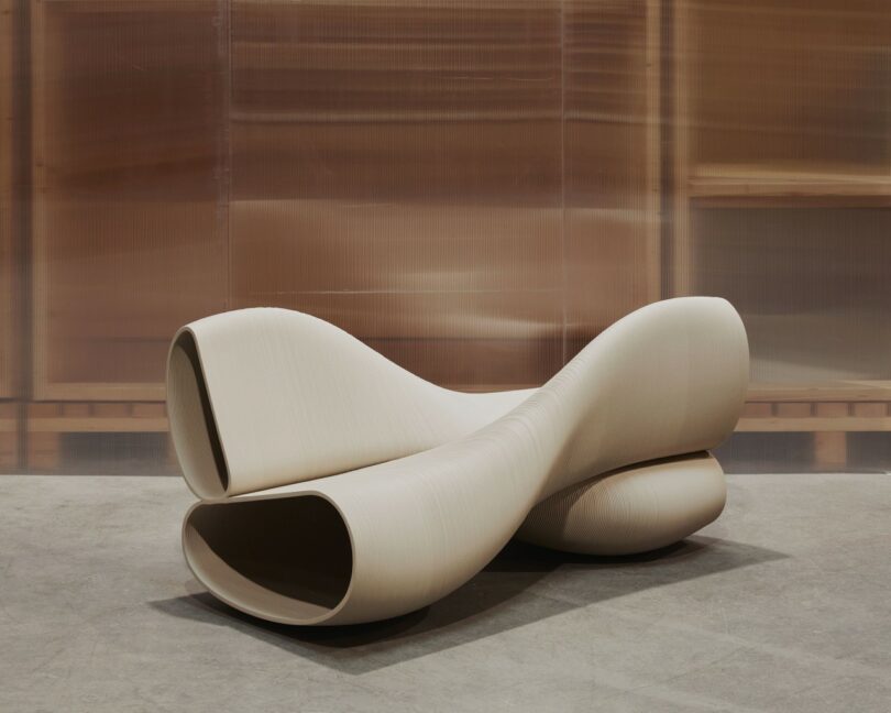 A modern, sculptural beige bench with smooth curves and hollowed ends sits on a concrete floor against a blurred brown background.