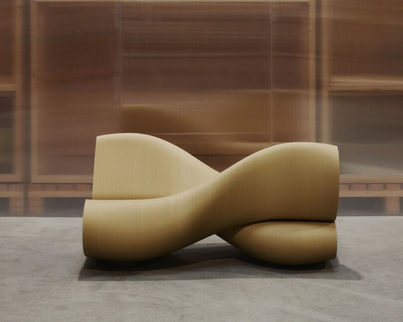 A sculptural, tan-colored bench with a twisting, wave-like design is positioned on a concrete floor
