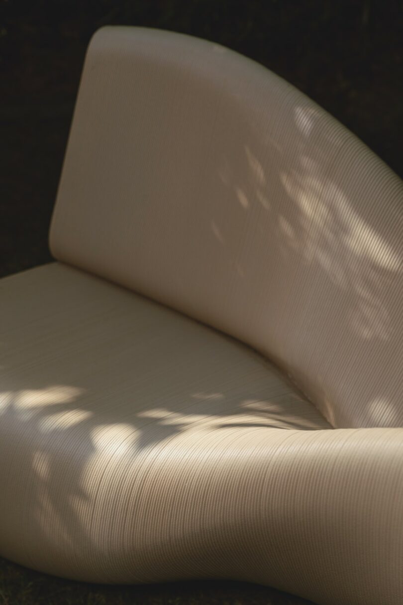 A modern, cream-colored, curved bench with a ribbed texture is partially shaded by dappled sunlight