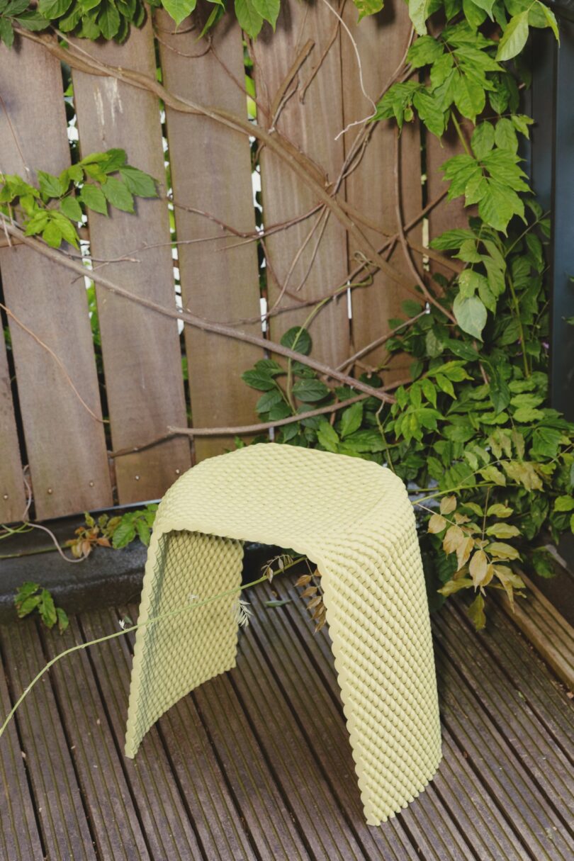 A textured, light yellow stool by The New Raw is placed on a wooden deck next to a wooden fence with green leafy vines climbing it.