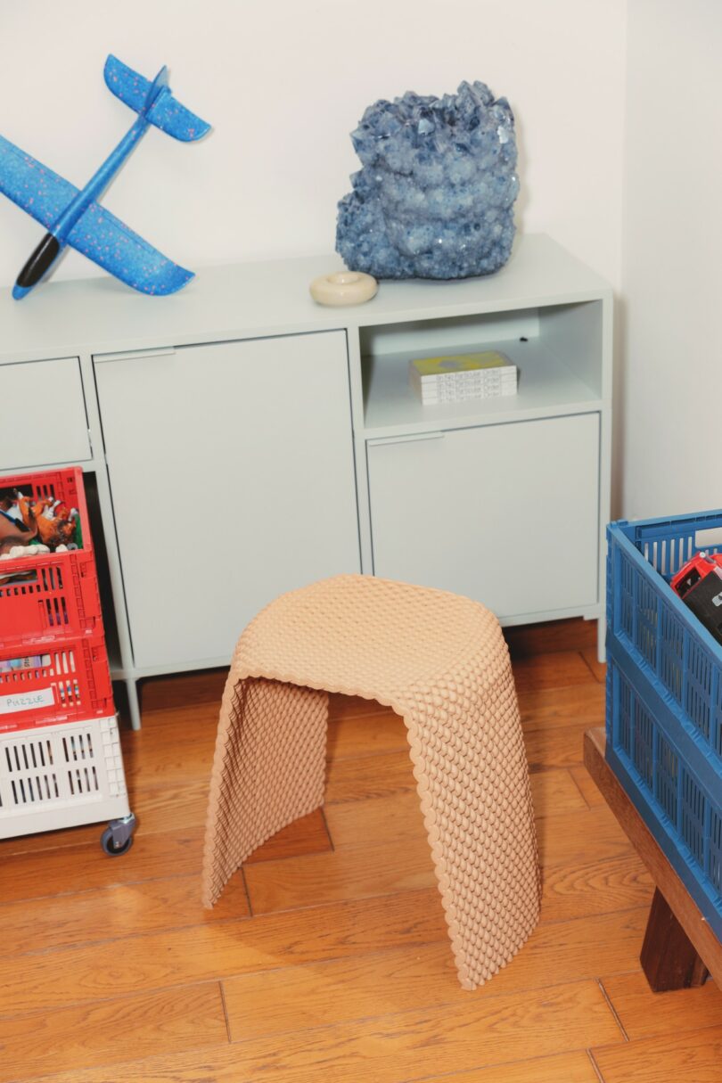 A textured beige stool sits on a wooden floor in front of a light gray cabinet, with a blue rock, toy plane, and plastic crates nearby