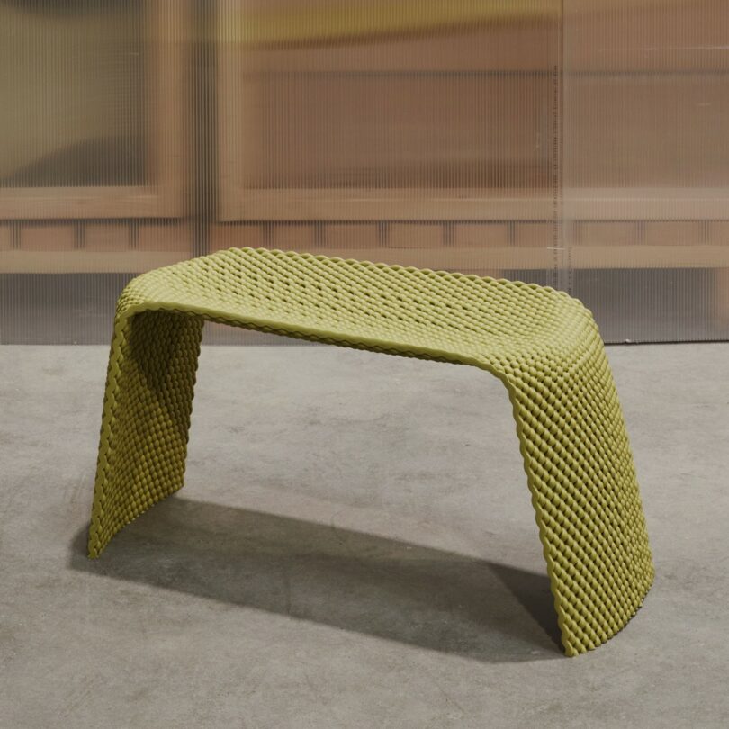 A yellow, textured bench with a curved, minimalist design sits on a concrete floor in front of a translucent wall