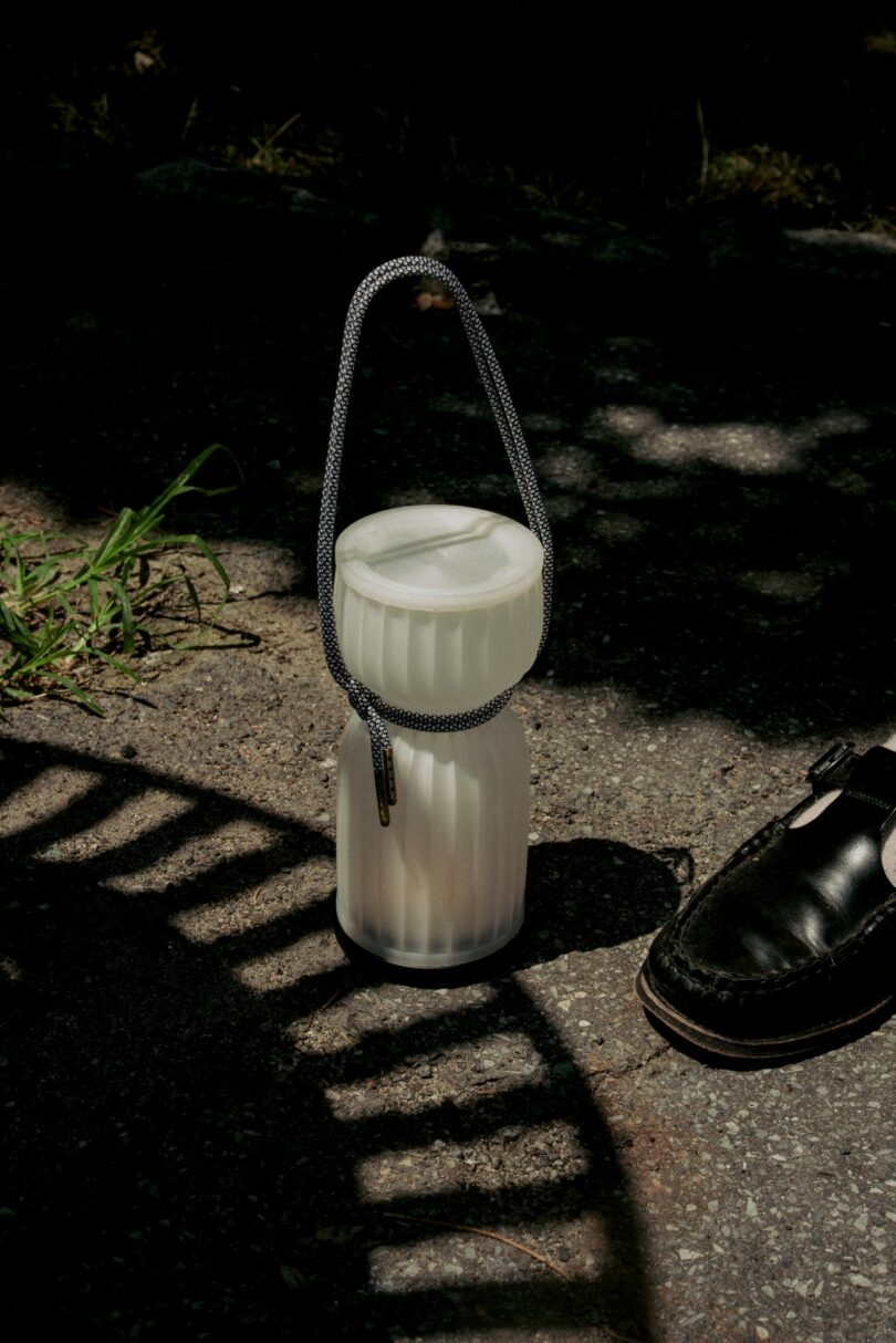 A white, ribbed container with a rope handle sits on pavement next to a black dress shoe, with some grass and shadows visible