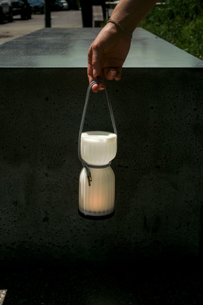 A hand holds a white, ribbed lantern by its cord in front of a concrete surface outdoors