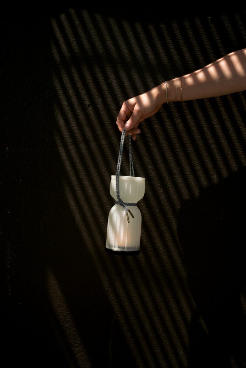 A hand holds a small white lantern by its strap against a black wall with diagonal light and shadow patterns