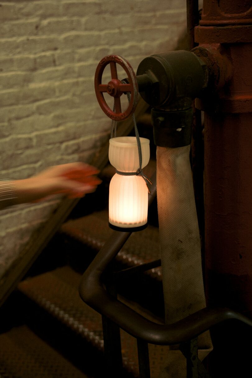 A white lantern hangs by a cord from a metal valve on a stairwell railing, with a blurred hand reaching toward it