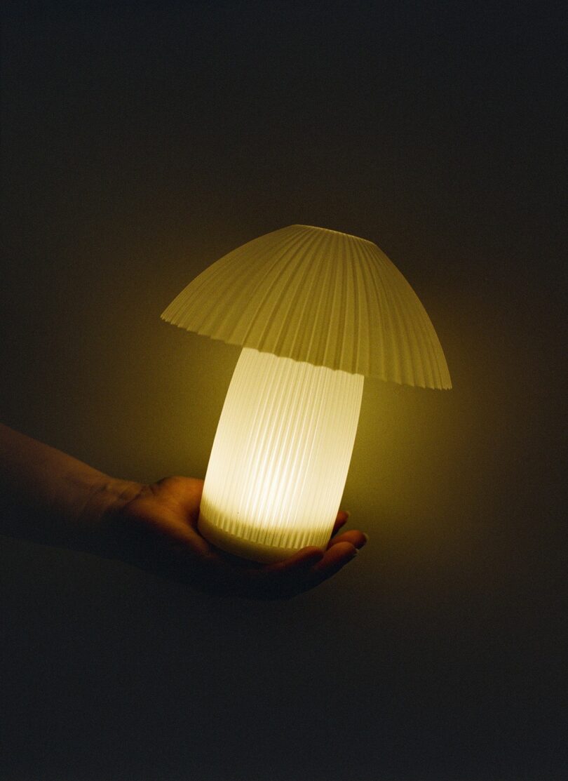 A hand holds a glowing, mushroom-shaped lamp with a ribbed, pleated shade against a dark background
