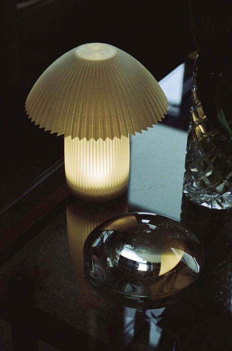 A pleated mushroom-shaped lamp emits soft light on a glass table next to a decorative clear glass object and a cut glass vase