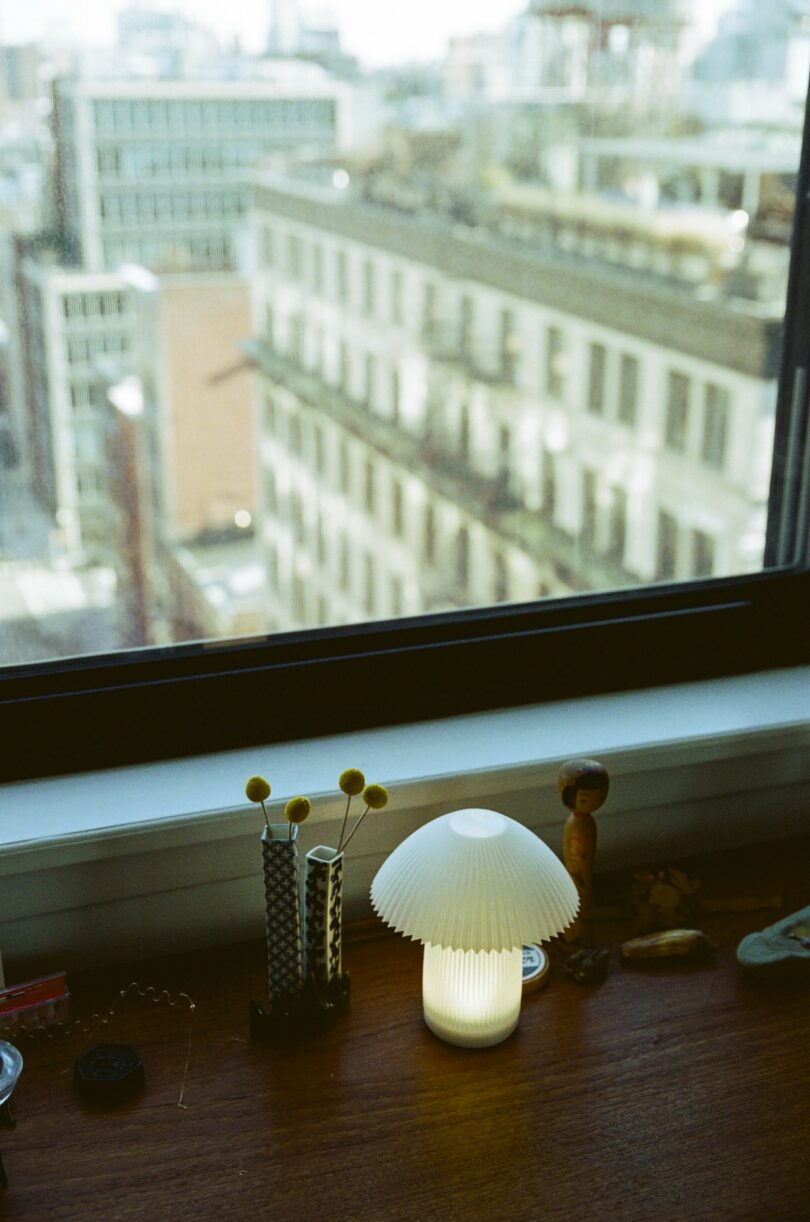 A lit white table lamp sits on a wooden windowsill with small decorative items, overlooking a cityscape through a large window