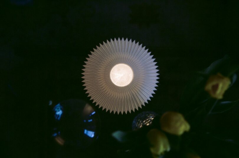 A white, circular, pleated lampshade is lit from within, photographed from above in a dimly lit room with yellow flowers visible in the bottom right corner